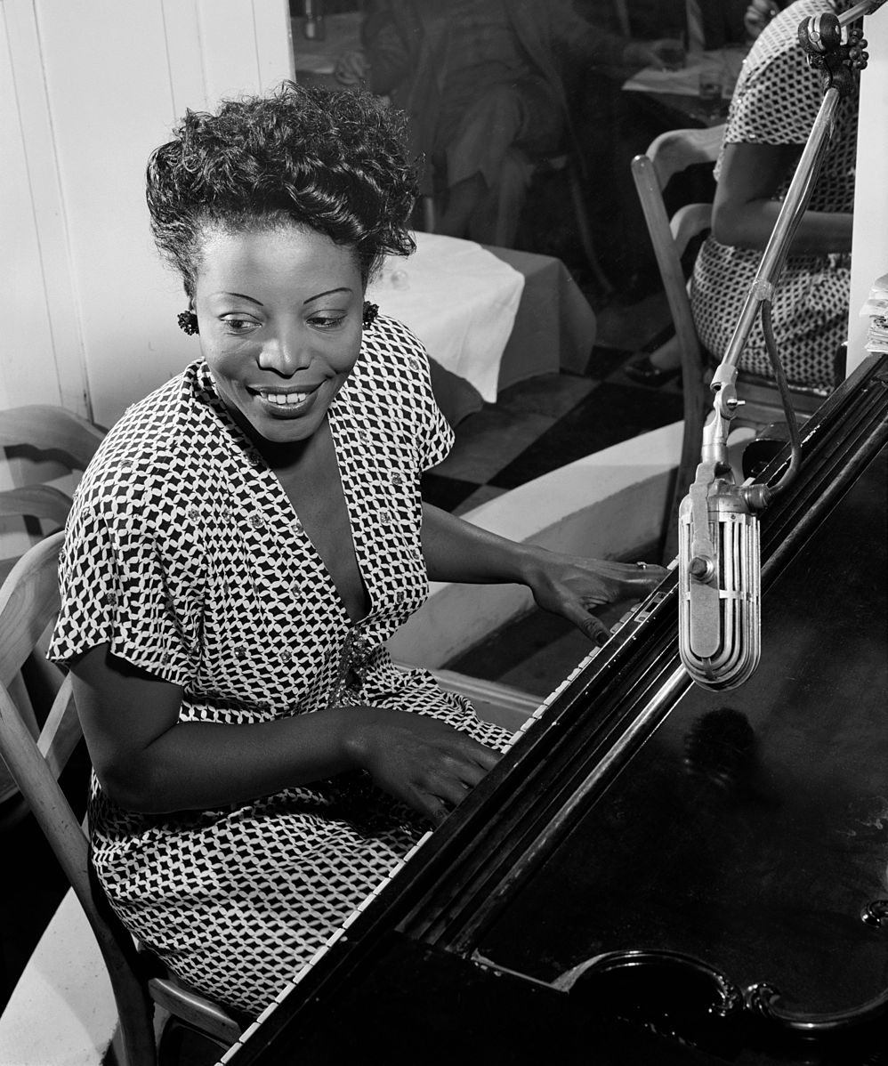 Black and white photo of woman with short curly hair in dotty dress playing piano with microphone in front.