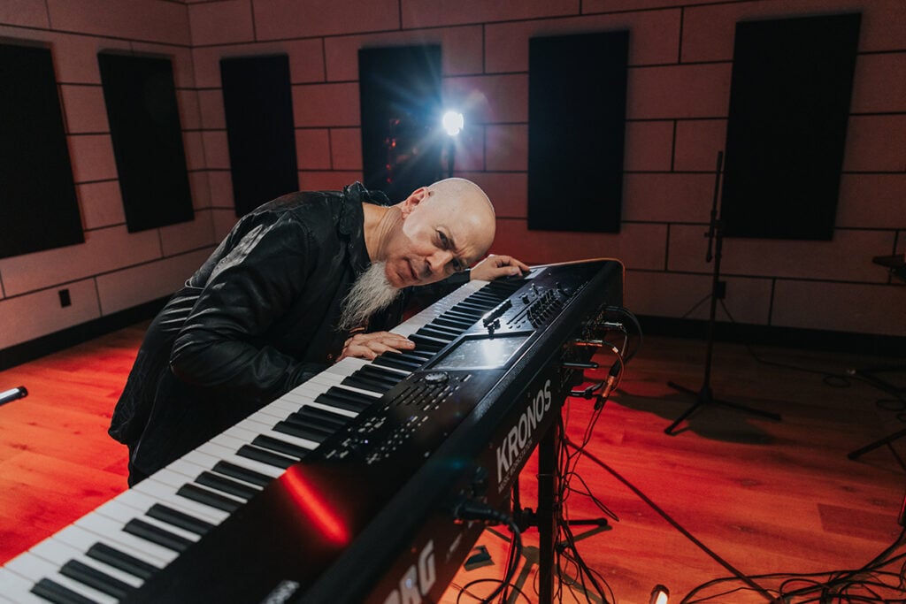 Man with goatee in leather jacket playing and leaning over keyboard in studio.