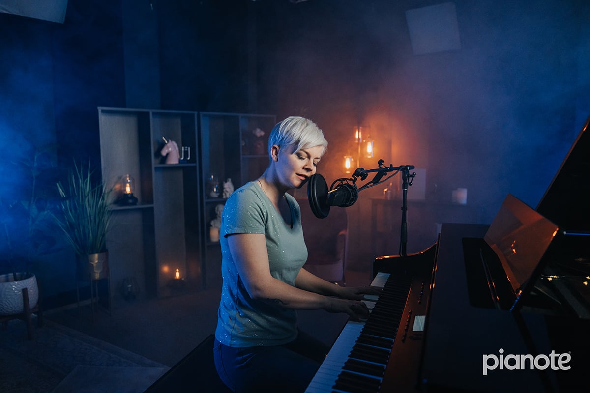 Woman with short platinum hair singing into a mic while playing piano in a dark blue lit studio.