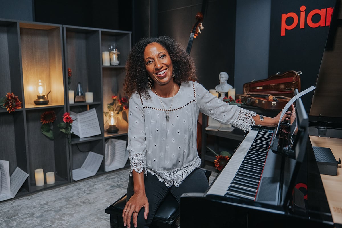Victoria Theodore (woman with curly hair in lacey top) sitting next to piano with one hand on music stand facing camera.